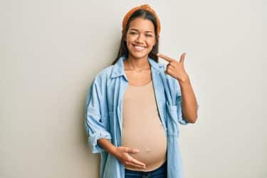 Beautiful hispanic woman expecting a baby, touching pregnant belly smiling cheerful showing and pointing with fingers teeth and mouth. dental health concept.